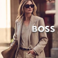 Eine Frau in einem beigen Blazer und mit Sonnenbrille, die eine Handtasche trägt, auf der das BOSS-Logo abgebildet ist.