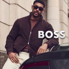 Ein Mann in einer braunen Jacke, mit Sonnenbrille und einer hellen Hose lehnt an einem Auto neben der Aufschrift „BOSS“.