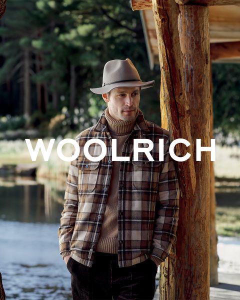 Ein Mann in einer karierten Jacke und mit Hut steht neben einer Holzhütte in der Nähe eines Teiches; über dem Bild ist der Schriftzug „WOOLRICH“ zu sehen.