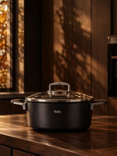 Auf einer hölzernen Küchenarbeitsplatte steht ein schwarzer Fissler-Topf mit Glasdeckel, während im Hintergrund warmes Sonnenlicht durch ein Fenster fällt.