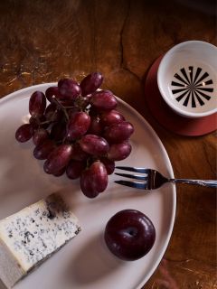 Ein Teller mit roten Trauben, eine Scheibe Blauschimmelkäse und eine Pflaume; außerdem sind eine Gabel und eine Tasse auf einem Holztisch zu sehen.