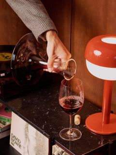 Eine Person schenkt Rotwein aus einer Karaffe in ein Glas neben einer roten Lampe ein, im Hintergrund sind ein David-Bowie-Album und ein Plattenspieler zu sehen.