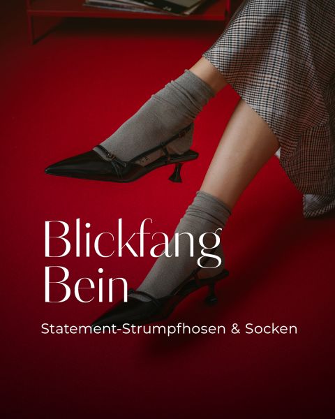 Eine Person mit grauen Socken, schwarzen Pumps und einem karierten Rock auf einem roten Boden. Text: „Blickfang Bein. Statement-Strumpfhosen & Socken.“.