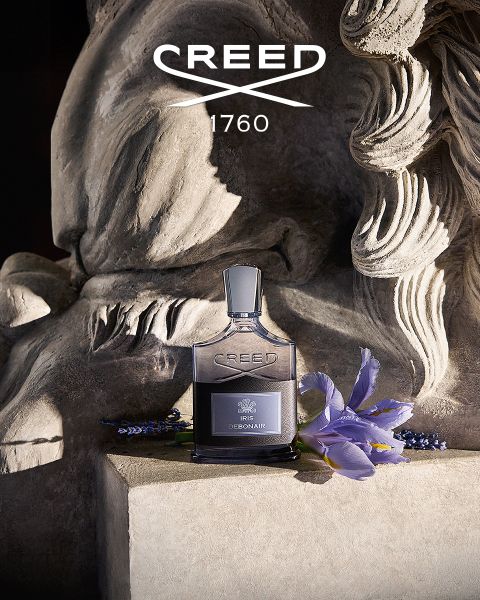 Neben einer Steinskulptur mit violetten Blumen steht ein Creed-Parfümflakon. Darüber steht in weißer Schrift „CREED 1760“.