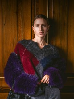 Eine Frau in einem flauschigen Pullover mit roten, blauen und violetten Streifen steht vor einer Holzwand, hält eine Tasche in der Hand und blickt in die Kamera.