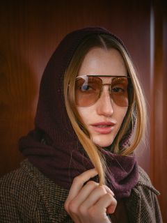 Eine Frau mit langen Haaren, die eine Pilotenbrille trägt, einen braunen Schal über dem Kopf hat und einen braunen Karomantel trägt, steht vor einer Holzwand.