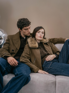 Ein Mann und eine Frau in olivfarbenen Jacken sitzen zusammen auf einem hellen Sofa.