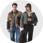 Zwei Models tragen Herbstoutfits: Der Mann trägt eine braune Jacke und Jeans, die Frau eine grüne Daunenjacke, ein gestreiftes Hemd, eine dunkle Hose und eine Pelzmütze.