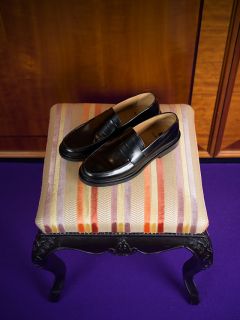 Ein Paar schwarze Leder-Loafer liegt auf einem gestreiften, gepolsterten Hocker mit verzierten schwarzen Beinen, der auf einem violetten Teppich steht.