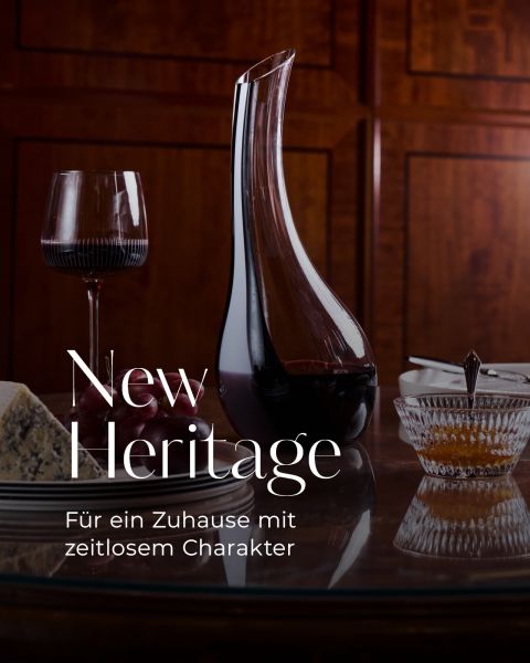 Eine Weinkaraffe, ein Glas Rotwein, Käse und Marmelade auf einem Holztisch. Der Text lautet: „New Heritage. Für ein Zuhause mit zeitlosem Charakter.“.
