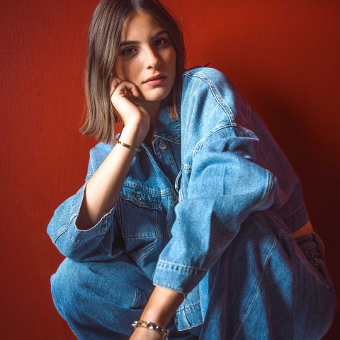 Eine Frau in Jeansjacke und Jeans sitzt vor einer leuchtend roten Wand, stützt ihr Kinn auf die Hand und blickt in die Kamera.