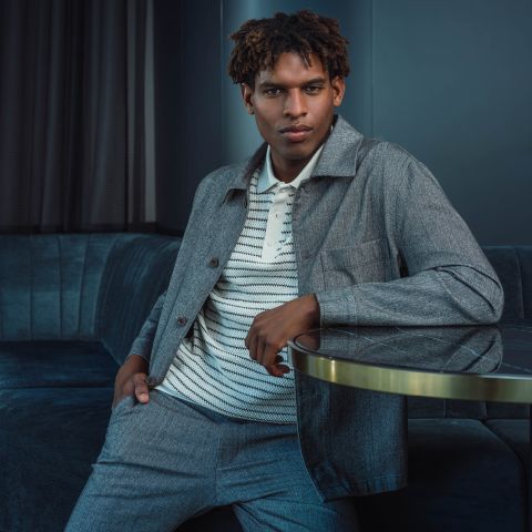 Ein Mann in einem grauen Anzug und einem gestreiften Hemd sitzt auf einem blauen Sofa, stützt einen Arm auf einen Glastisch und blickt selbstbewusst in die Kamera.