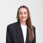 Eine Frau mit langen braunen Haaren, die einen schwarzen Blazer und ein weißes Oberteil trägt, lächelt leicht und steht vor einem schlichten, hellen Hintergrund.