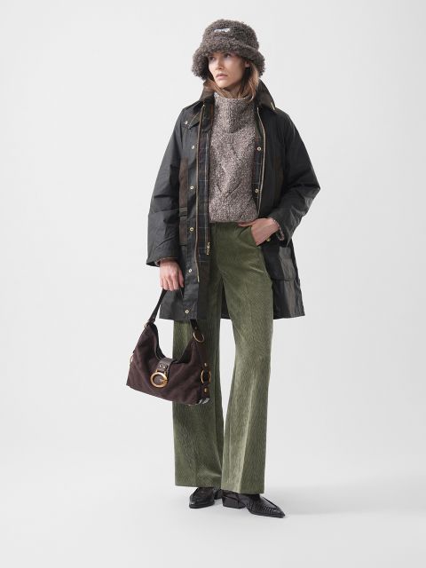 Eine Frau mit einer Pelzmütze, einer dunklen Jacke, einem braunen Pullover, einer grünen Cordhose und schwarzen Schuhen, die eine braune Handtasche in der Hand hält und vor einem schlichten Hintergrund steht.