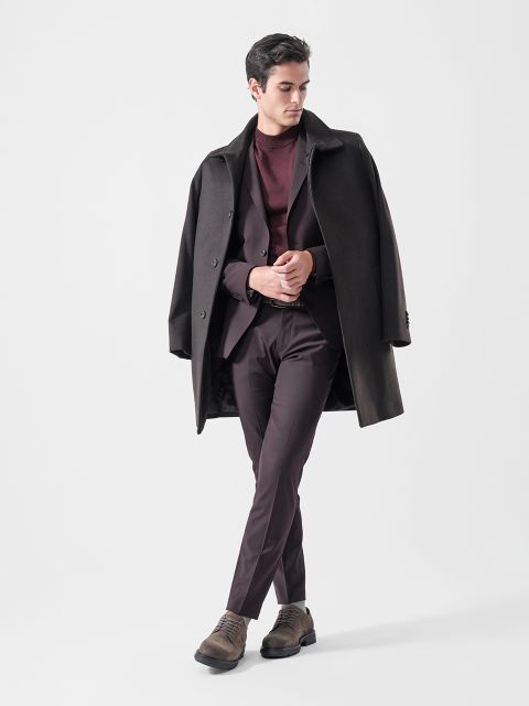 Ein Mann, der einen dunklen Mantel über die Schultern gelegt hat, einen burgunderroten Anzug, einen burgunderroten Pullover und braune Schuhe trägt und vor einem schlichten, hellen Hintergrund steht.