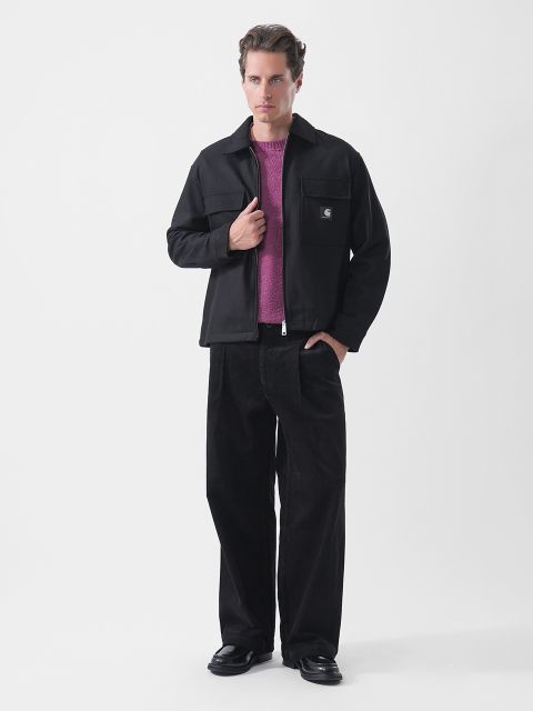 Ein Mann posiert in einer schwarzen Jacke über einem lila Pullover, einer schwarzen Hose mit weitem Bein und schwarzen Schuhen vor einem schlichten weißen Hintergrund.