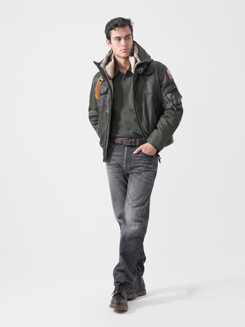 Ein Mann, der eine dunkelgrüne Jacke, ein grünes Hemd und Jeans trägt, steht mit den Händen in den Taschen vor einem schlichten weißen Hintergrund.