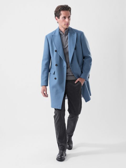 Ein Mann in einem hellblauen Zweireiher, einem grauen Hemd, einer dunklen Hose und schwarzen Stiefeln, der selbstbewusst vor einem schlichten weißen Hintergrund geht.
