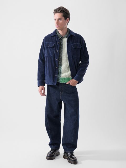 Ein Mann, der eine marineblaue Jacke, einen grün-weißen Pullover, eine weite dunkle Jeans und schwarze Schuhe trägt, steht vor einem schlichten weißen Hintergrund.