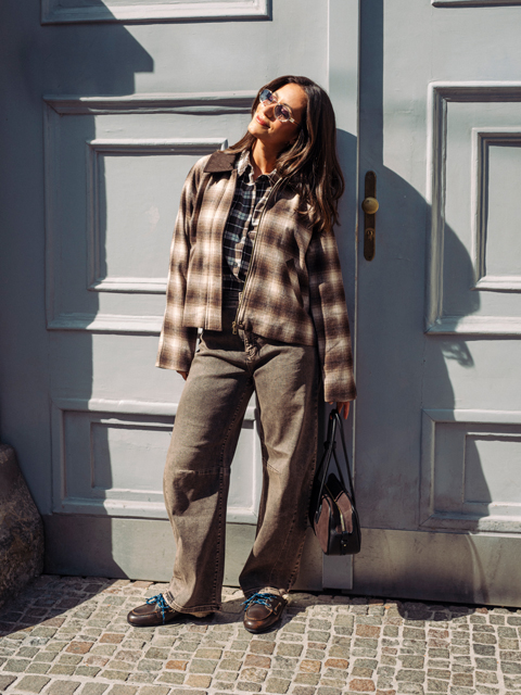 Eine Frau, die eine karierte Jacke, Jeans und Slipper trägt, lehnt an einer blauen Tür.