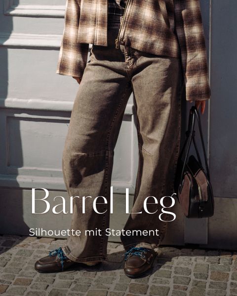Eine Person, die eine karierte Jacke, Jeans mit weitem Bein und braune Schuhe trägt, steht neben einer Tür, über der der Schriftzug „Barrel Leg Silhouette mit Statement“ zu lesen ist.