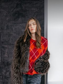 Eine Frau trägt einen rot-orangefarbenen Argyle-Pullover unter einer dunklen Kunstfelljacke.