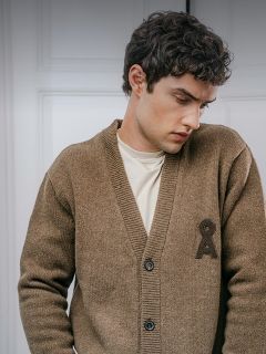 Ein Mann trägt eine braune Strickjacke mit Knöpfen und einem dunklen, aufgestickten Emblem auf der Brust über einem cremefarbenen T-Shirt.