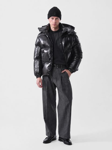 Ein Mann, der eine schwarze Mütze, eine glänzende schwarze Daunenjacke, ein schwarzes Hemd, eine graue Hose und schwarze Slipper trägt.
