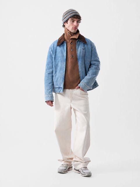 Ein Mann, der eine blaue Jeansjacke über einem braunen Pullover mit Knöpfen, eine cremefarbene Hose und eine Mütze trägt.