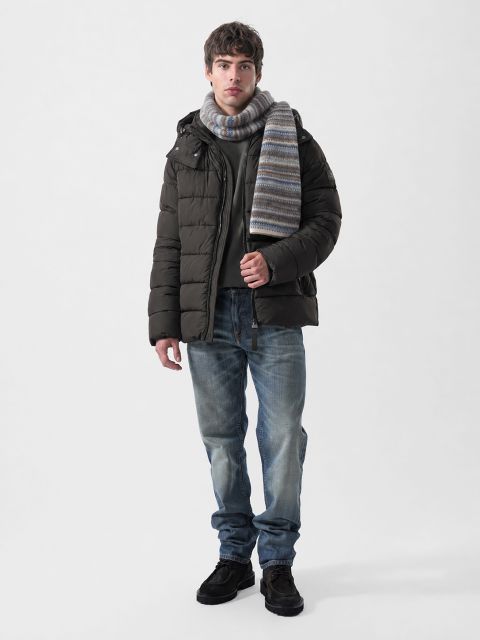Ein Mann, der eine dunkelbraune Daunenjacke, einen grauen Pullover, einen gestreiften Schal, blaue Jeans und dunkle Schuhe trägt.