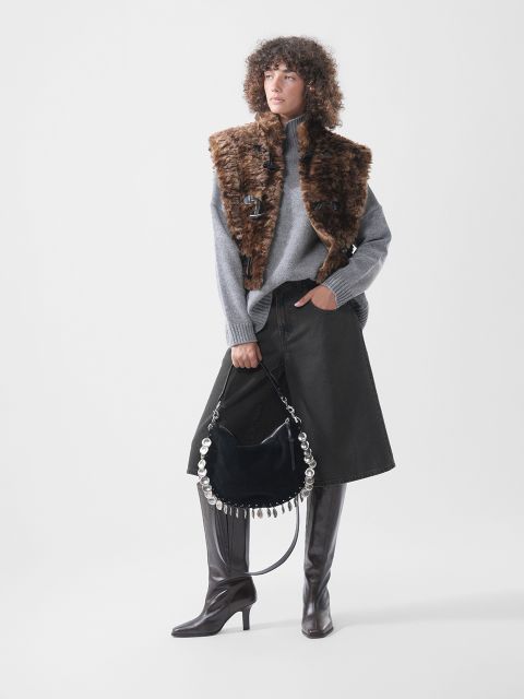 Ein Model trägt einen grauen Rollkragenpullover, eine braune Kunstfellweste und einen schwarzen Jeansrock; dazu hält sie eine schwarze Handtasche mit silbernen, runden Beschlägen und trägt hohe braune Stiefel.