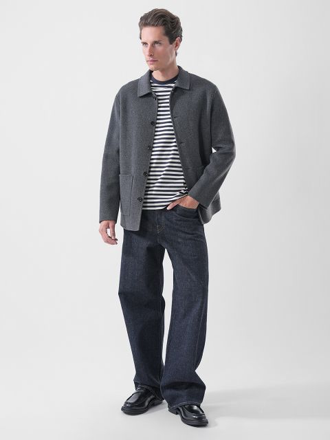 Ein Mann, der eine graue Jacke über einem marineblau-weiß gestreiften T-Shirt, dunkelblaue Jeans und schwarze Slipper trägt.