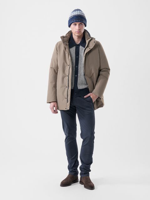 Ein Mann, der einen hellbraunen Parka über einem gemusterten Hemdpullover trägt, dazu eine dunkle Hose, braune Stiefel und eine gestreifte Strickmütze.