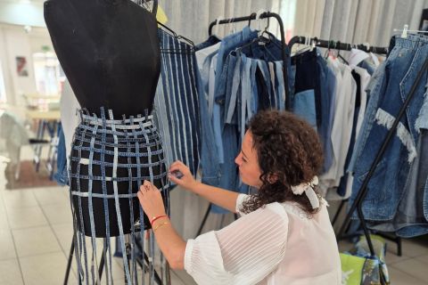 Eine Frau kniet in einem Atelier, hinter ihr stehen Kleiderständer mit Jeanskleidung, und befestigt Jeansstreifen an einer Schneiderpuppe.